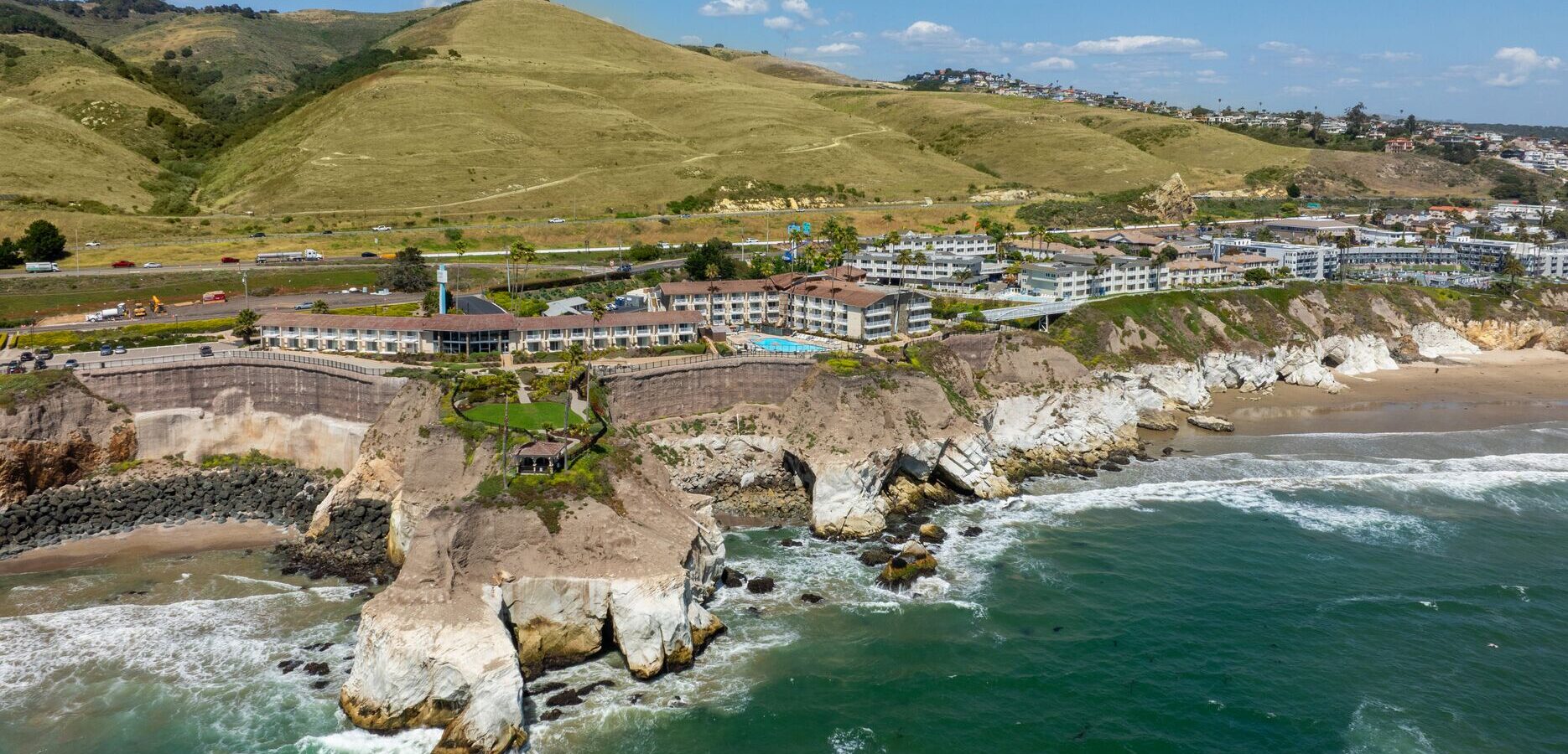 Aerial view of a coastal resort with buildings atop rocky cliffs, overlooking the ocean. Rolling green hills and a highway are visible in the background under a clear blue sky.