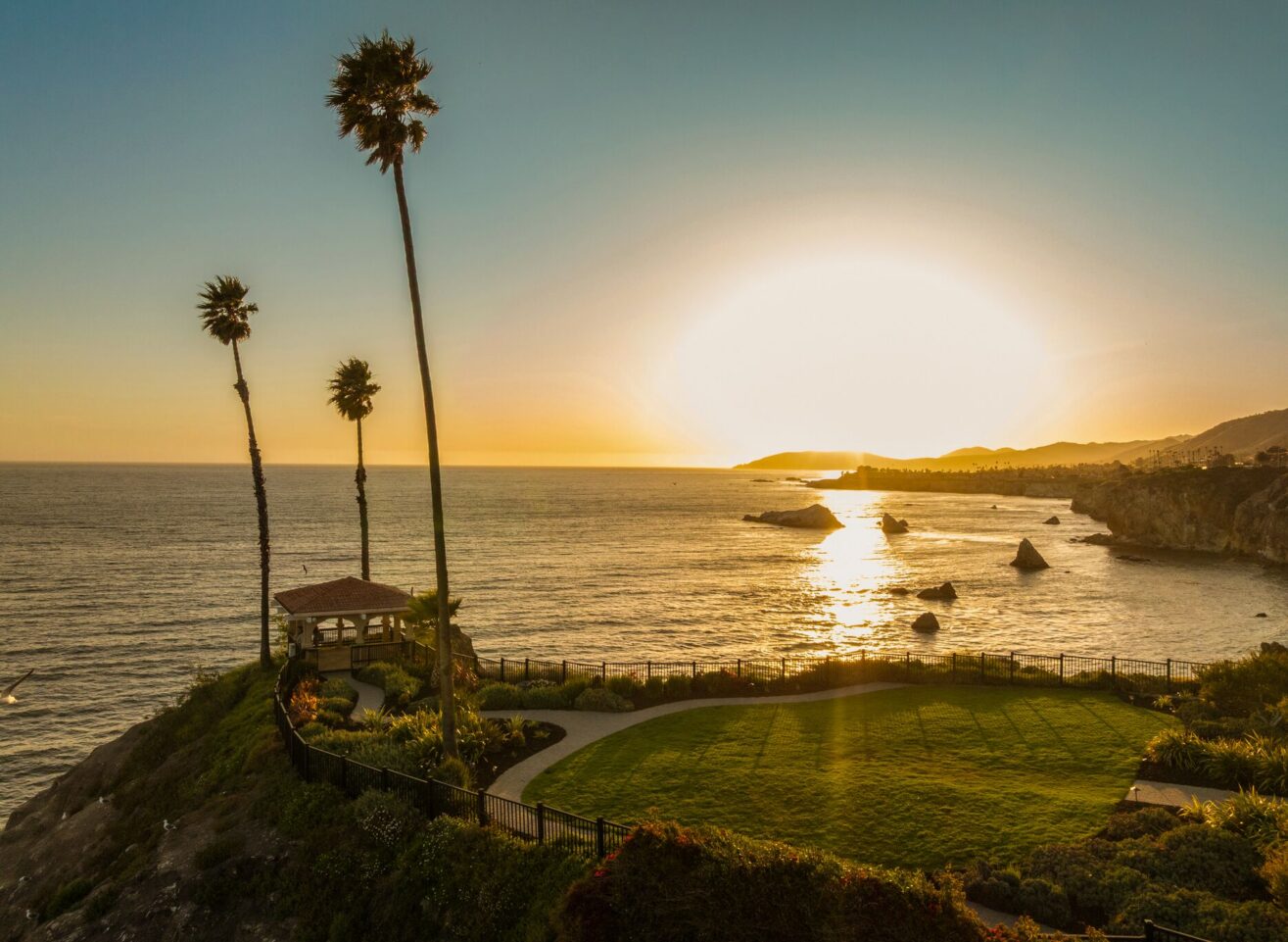 A coastal landscape at sunset, showing a grassy area with a small gazebo, tall palm trees, and the ocean reflecting the golden sun near the horizon. Rocky cliffs and distant hills frame the shoreline.