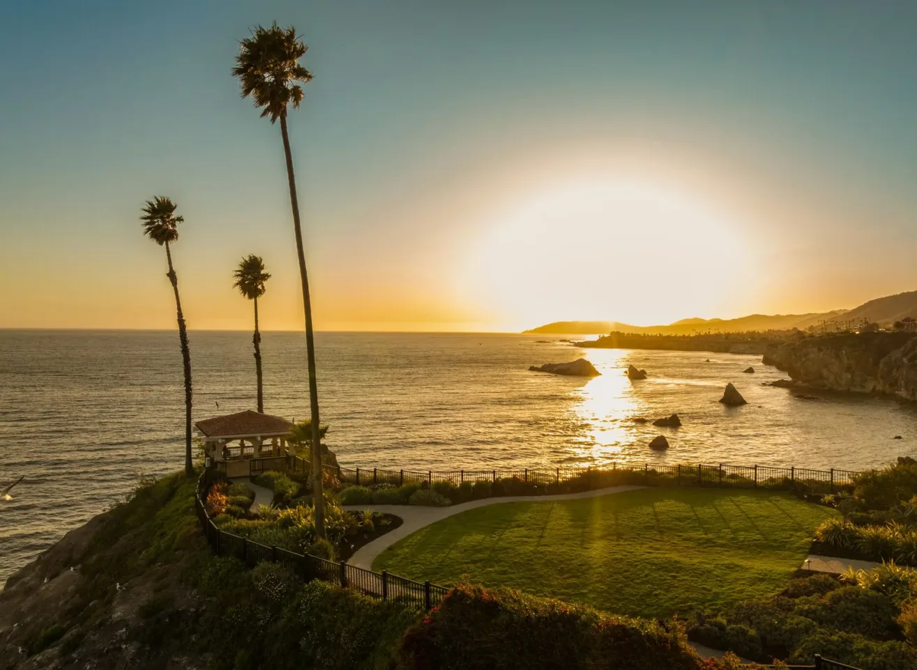 A coastal landscape at sunset, showing a grassy area with a small gazebo, tall palm trees, and the ocean reflecting the golden sun near the horizon. Rocky cliffs and distant hills frame the shoreline.