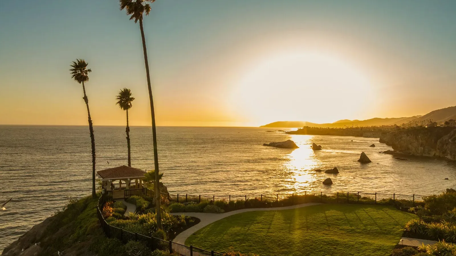 A coastal landscape at sunset, showing a grassy area with a small gazebo, tall palm trees, and the ocean reflecting the golden sun near the horizon. Rocky cliffs and distant hills frame the shoreline.