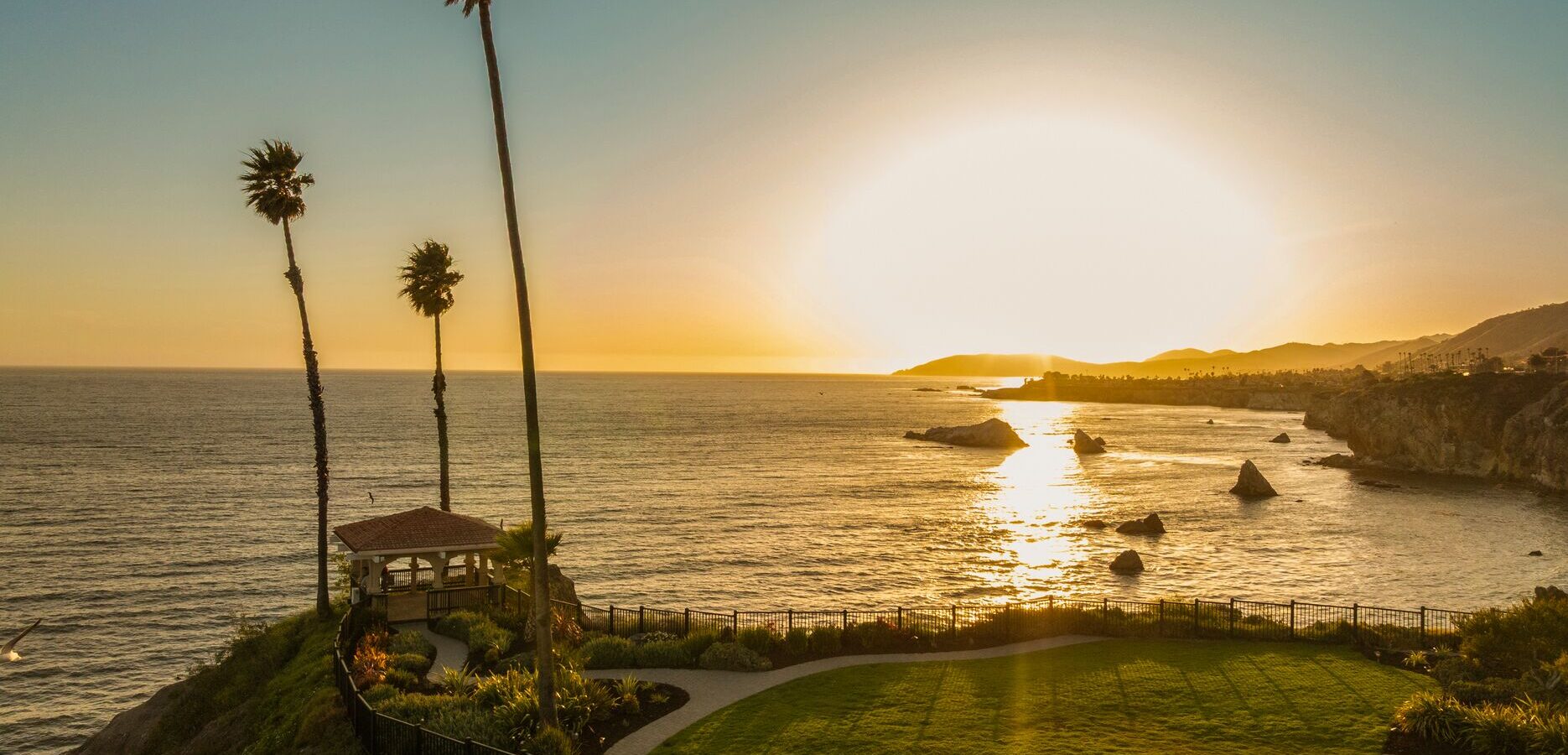 A coastal landscape at sunset, showing a grassy area with a small gazebo, tall palm trees, and the ocean reflecting the golden sun near the horizon. Rocky cliffs and distant hills frame the shoreline.
