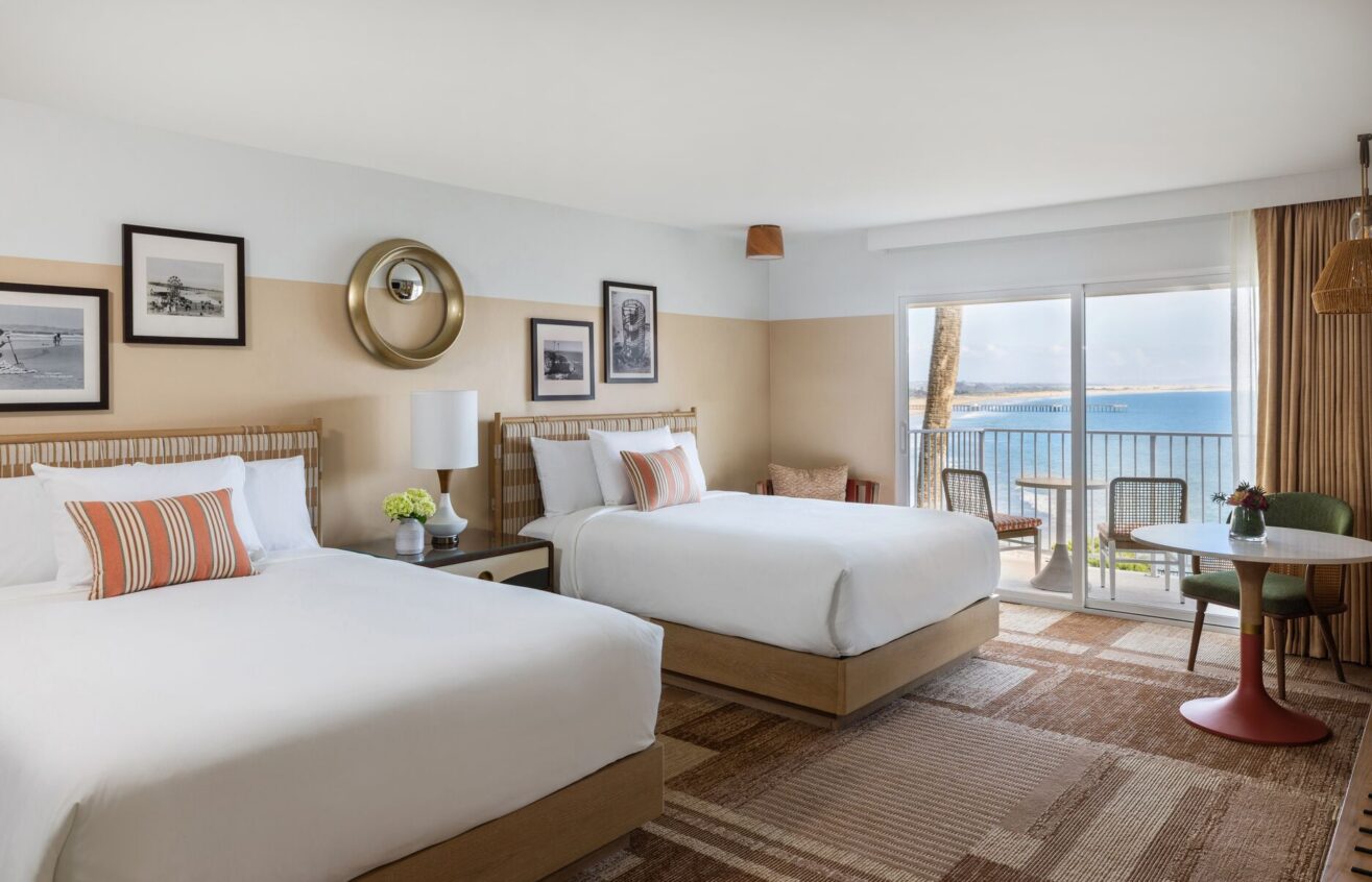 Bright hotel room with two double beds, striped accent pillows, framed art on the walls, a lamp, a small round table with chairs, and a balcony overlooking the ocean under a clear sky.