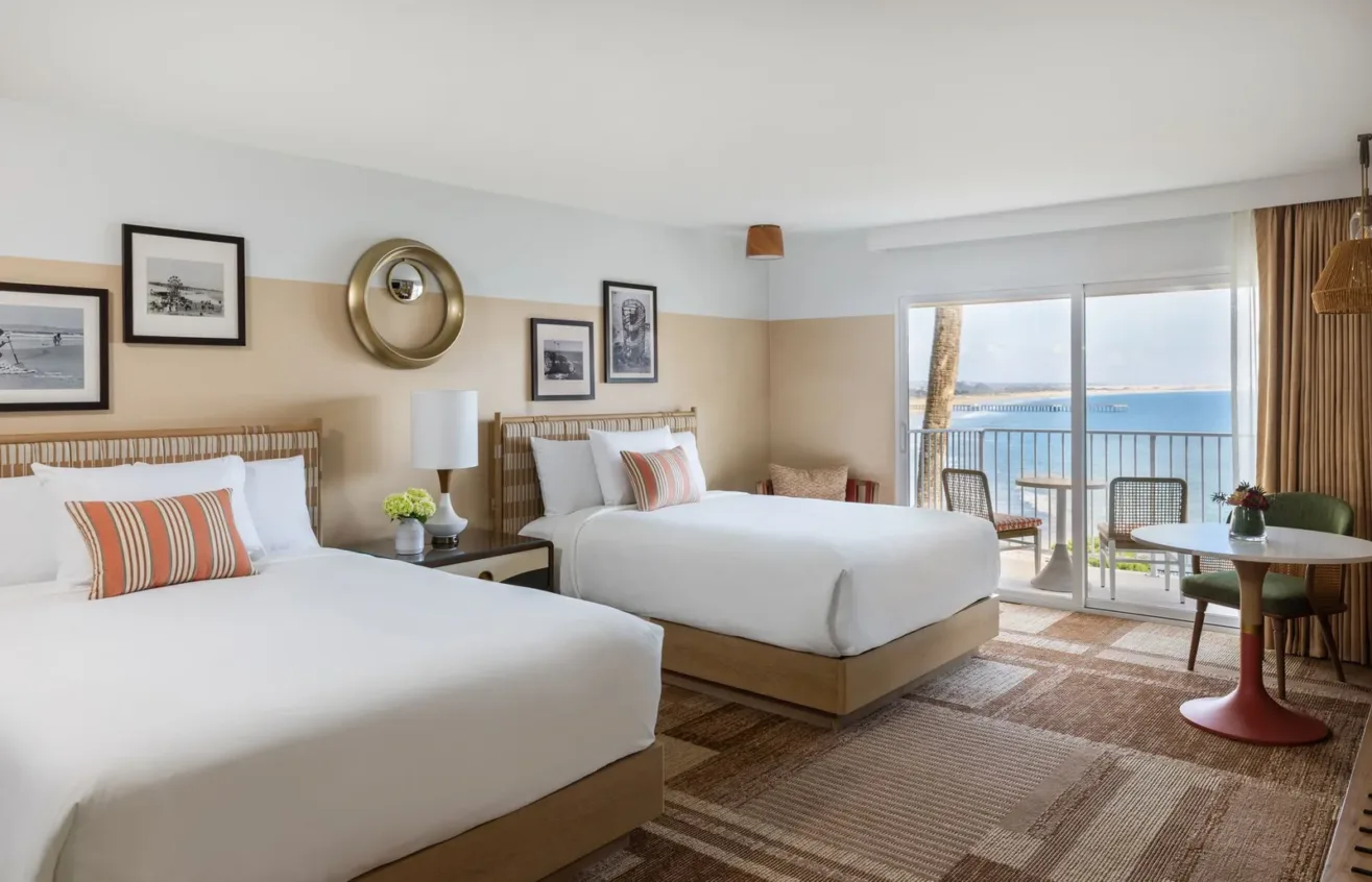 Bright hotel room with two double beds, striped accent pillows, framed art on the walls, a lamp, a small round table with chairs, and a balcony overlooking the ocean under a clear sky.