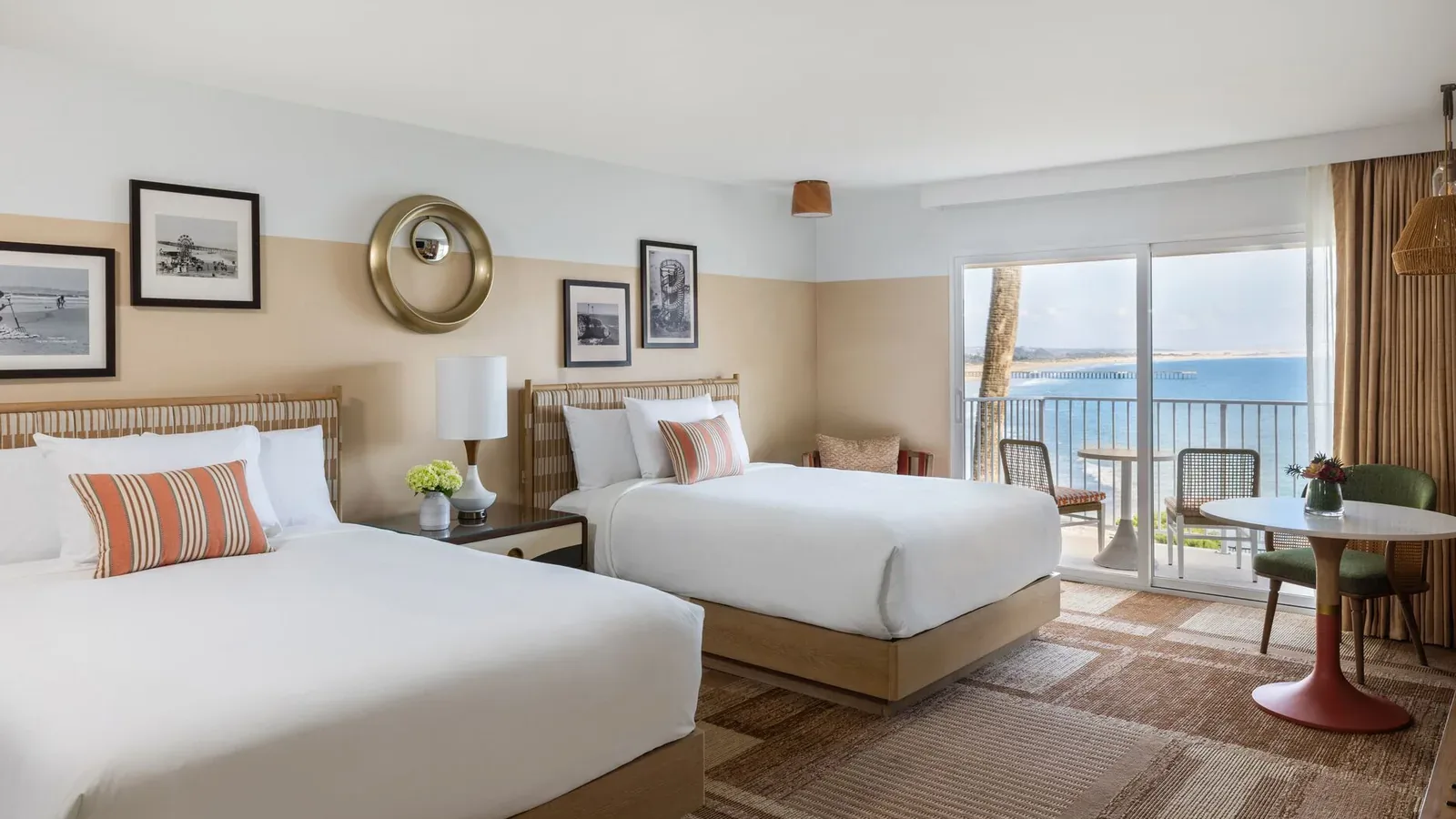 Bright hotel room with two double beds, striped accent pillows, framed art on the walls, a lamp, a small round table with chairs, and a balcony overlooking the ocean under a clear sky.