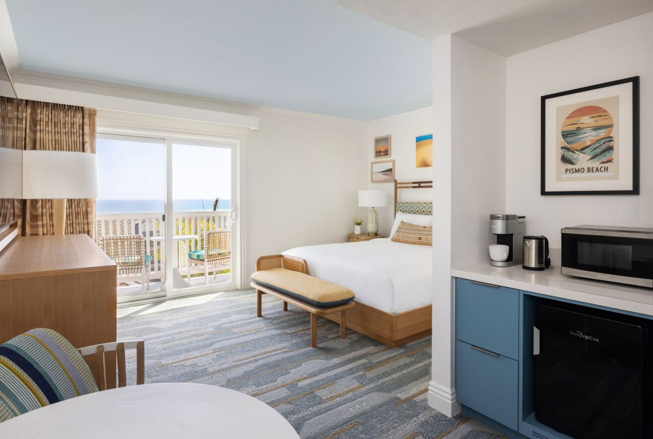 A bright, modern hotel room with a king bed, blue accents, a small table and chairs, a microwave, coffee maker, mini fridge, patio doors, and a balcony with an ocean view. Beach-themed artwork decorates the walls.