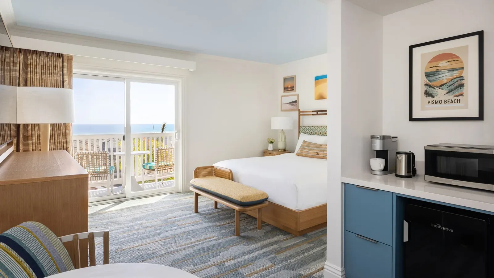 A bright, modern hotel room with a king bed, blue accents, a small table and chairs, a microwave, coffee maker, mini fridge, patio doors, and a balcony with an ocean view. Beach-themed artwork decorates the walls.