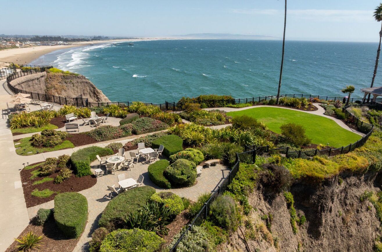 A landscaped garden with patios and seating overlooks the ocean from a cliff, with a sandy beach and distant horizon visible under a clear blue sky.