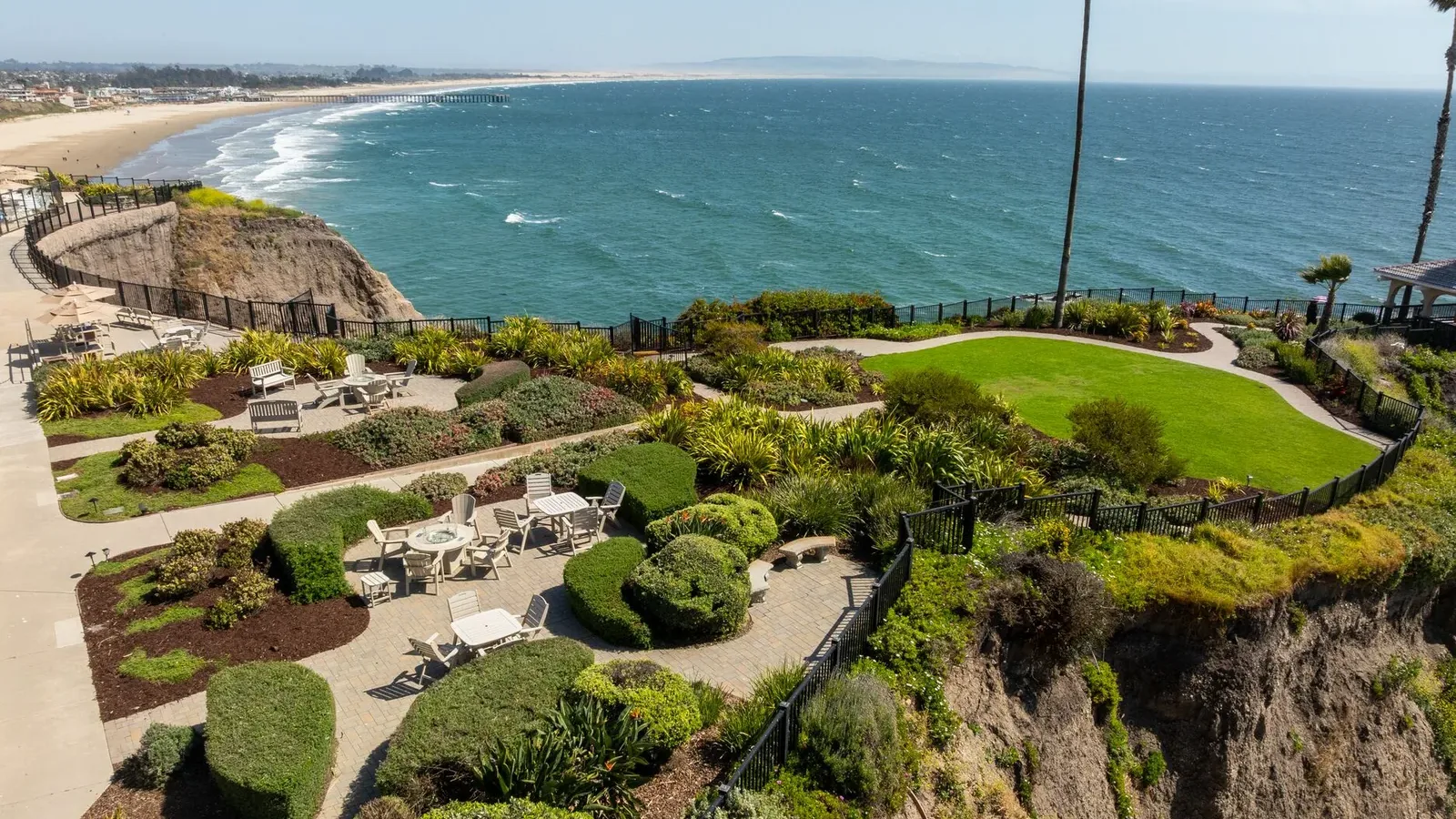 A landscaped garden with patios and seating overlooks the ocean from a cliff, with a sandy beach and distant horizon visible under a clear blue sky.
