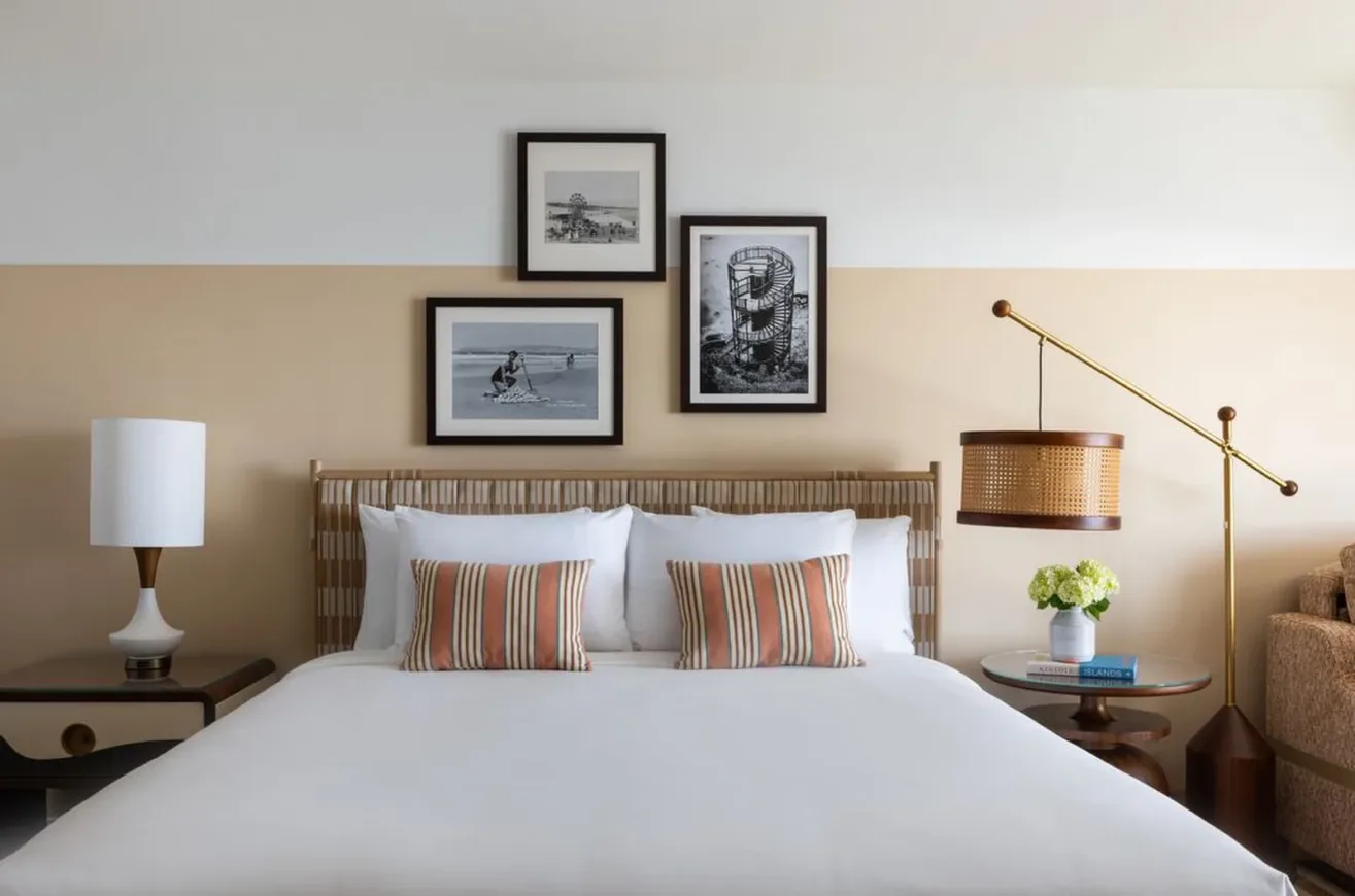 A neatly made bed with white linens and striped pillows is flanked by two side tables, lamps, and a modern floor lamp. Three black-and-white photos hang on the wall above the headboard.