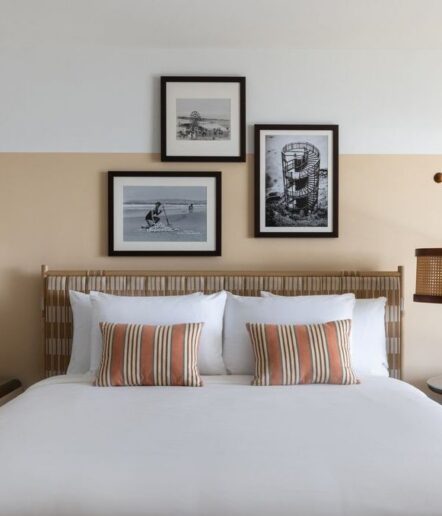 A neatly made bed with white linens and two striped orange and white pillows is set against a beige and white wall. Above the bed are three black-and-white framed photographs arranged in a cluster.