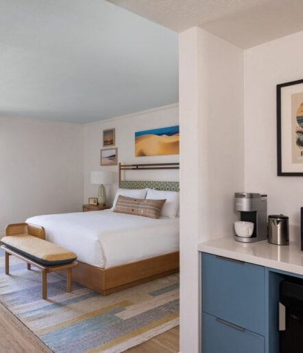 A modern hotel room with a neatly made bed, a wooden bench, colorful rug, wall art, and a kitchenette area with a coffee maker and mugs on the counter. The decor is minimal and inviting.