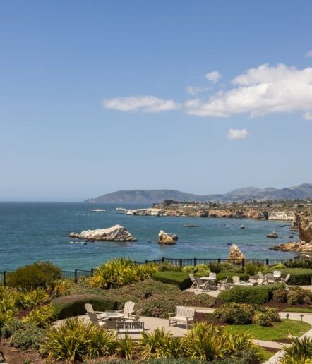 A scenic coastal landscape with neatly arranged lounge chairs on a landscaped garden overlooking the ocean, rocky shoreline, and distant hills under a blue sky with scattered clouds.