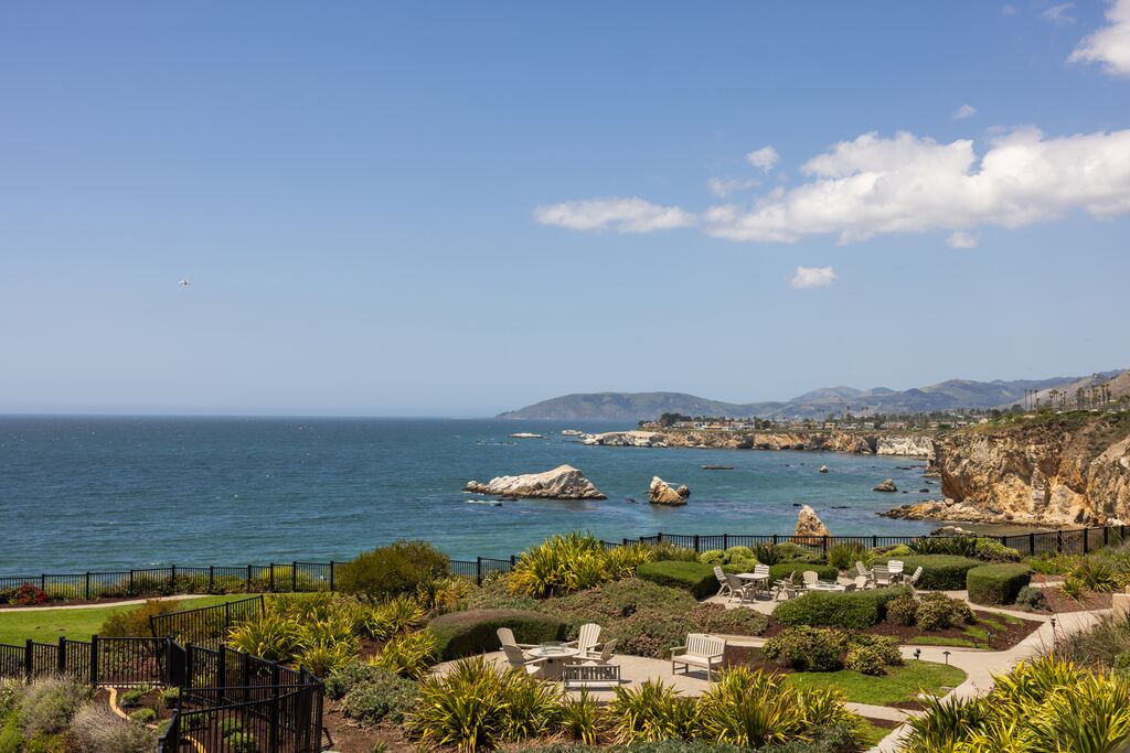 A scenic coastal view with landscaped gardens, outdoor seating, rocky cliffs, and blue ocean water under a partly cloudy sky. Hills are visible in the background.