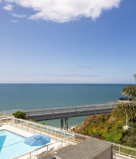 A rooftop view overlooking a swimming pool with a blue umbrella, a wooden pier extending over the ocean, and a clear, sunny sky with a few clouds.