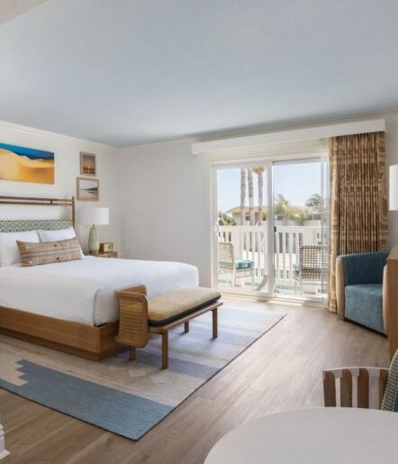 A bright hotel room with a large bed, wooden furniture, blue armchair, patterned rug, and sliding glass doors leading to a balcony with outdoor chairs and palm trees visible outside.