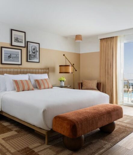 A modern hotel room features a large bed with striped pillows, framed art above the headboard, a brown padded bench, a woven rug, a chair, and glass doors opening to a balcony with an ocean view.