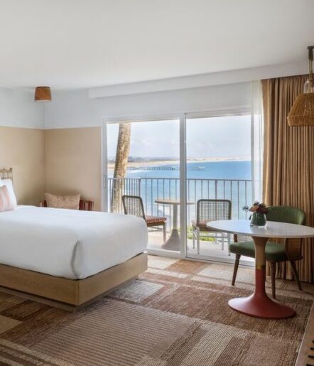 A bright hotel room with a large bed, woven rugs, a small round table with two chairs, and sliding glass doors opening to a balcony overlooking the ocean.