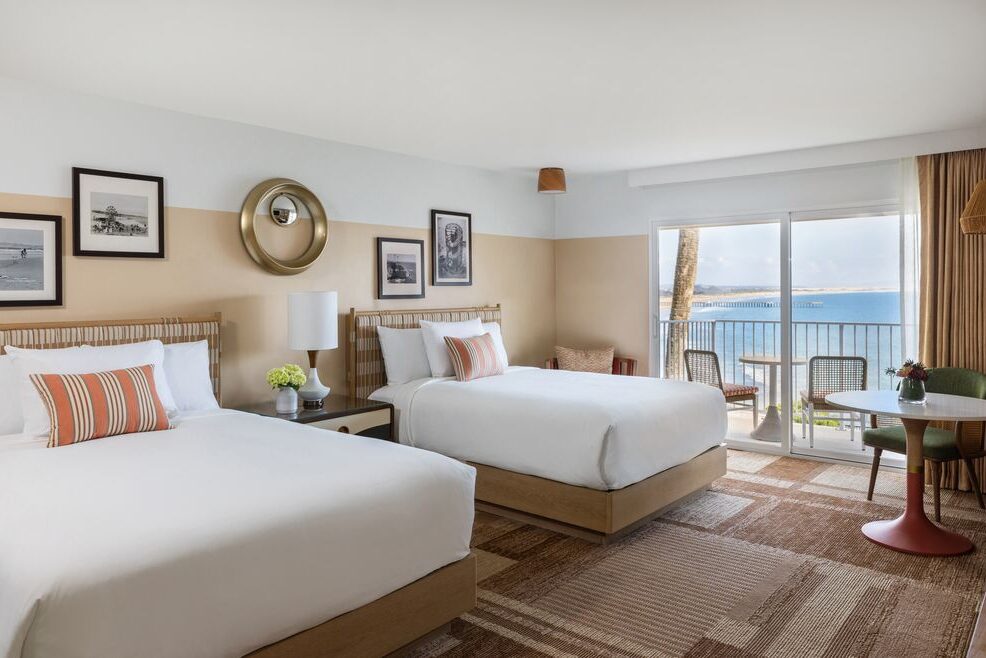 A hotel room with two double beds, striped pillows, wall art, a lamp, a small round table with two chairs, and a sliding glass door leading to a balcony with an ocean view.