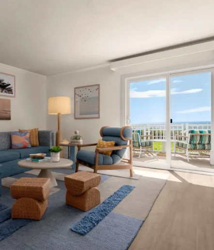 Bright living room with modern furniture, textured rug, and large sliding glass doors opening to a balcony with a scenic ocean view under a blue sky. Decor includes art, plants, and cozy seating.