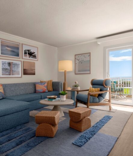 A bright living room with a blue sectional sofa, accent chair, round coffee table, woven stools, and a floor lamp. Ocean-themed art decorates the walls, and sunlight streams in through glass doors leading to a balcony.