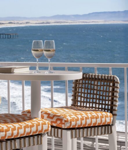 A round table with two glasses of white wine, two books, and two chairs with orange and white cushions overlooks the ocean from a balcony, with waves and distant hills in the background.