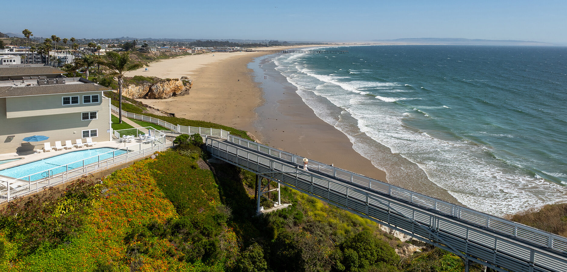 A coastal scene showing a sandy beach with gentle waves, a house with a swimming pool on a cliff, and a bridge extending from the house toward the beach under a clear blue sky.
