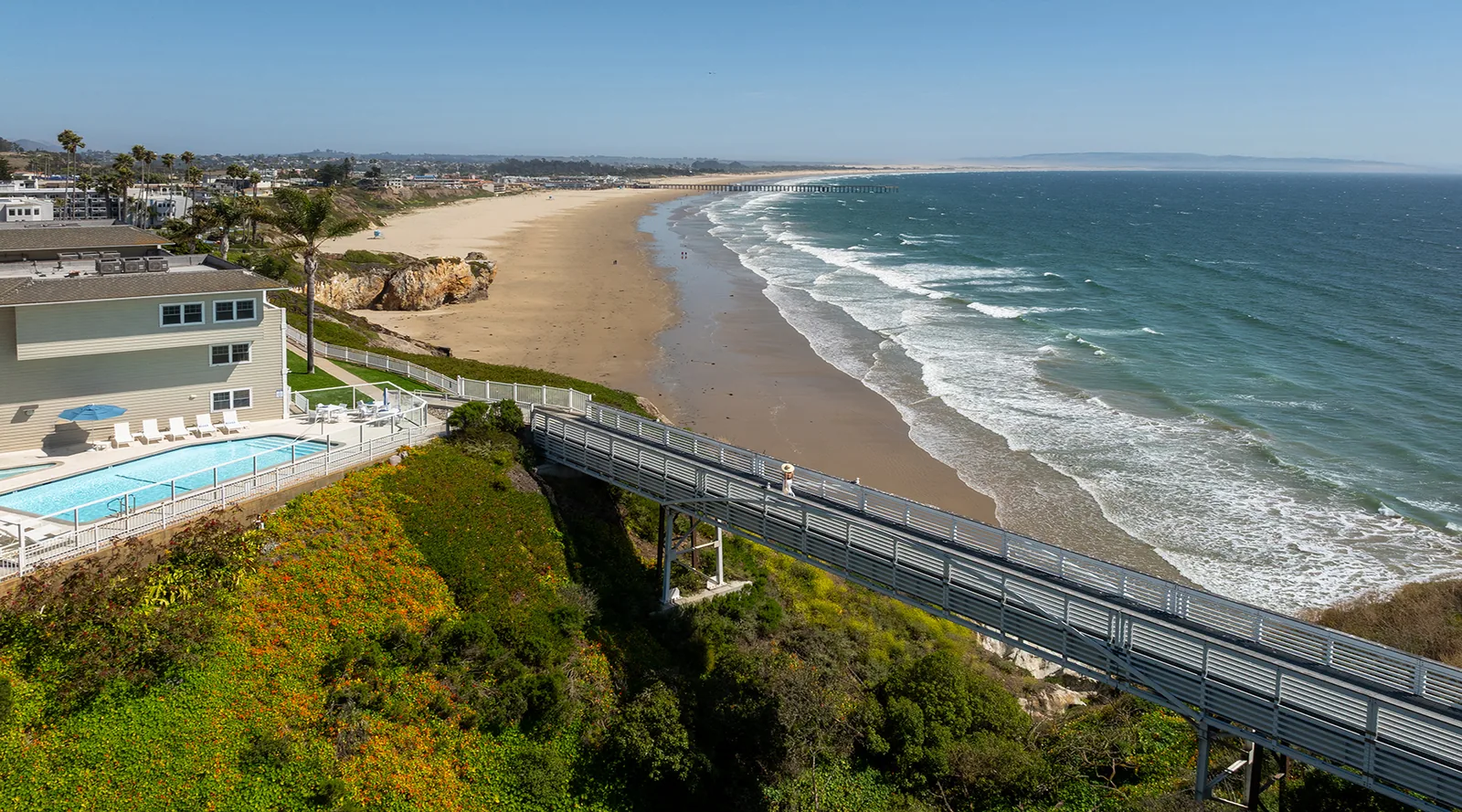 A coastal scene showing a sandy beach with gentle waves, a house with a swimming pool on a cliff, and a bridge extending from the house toward the beach under a clear blue sky.