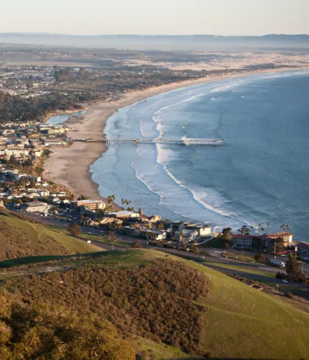 Aerial view of a coastal city with a long pier extending into the ocean, wide sandy beach, gentle waves, and green hills in the foreground under clear skies.
