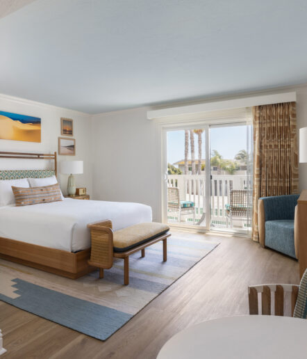 A bright hotel room with a large bed, a bench, modern décor, wooden furniture, a blue and beige rug, and a sliding glass door leading to a balcony with palm trees and a pool visible outside.