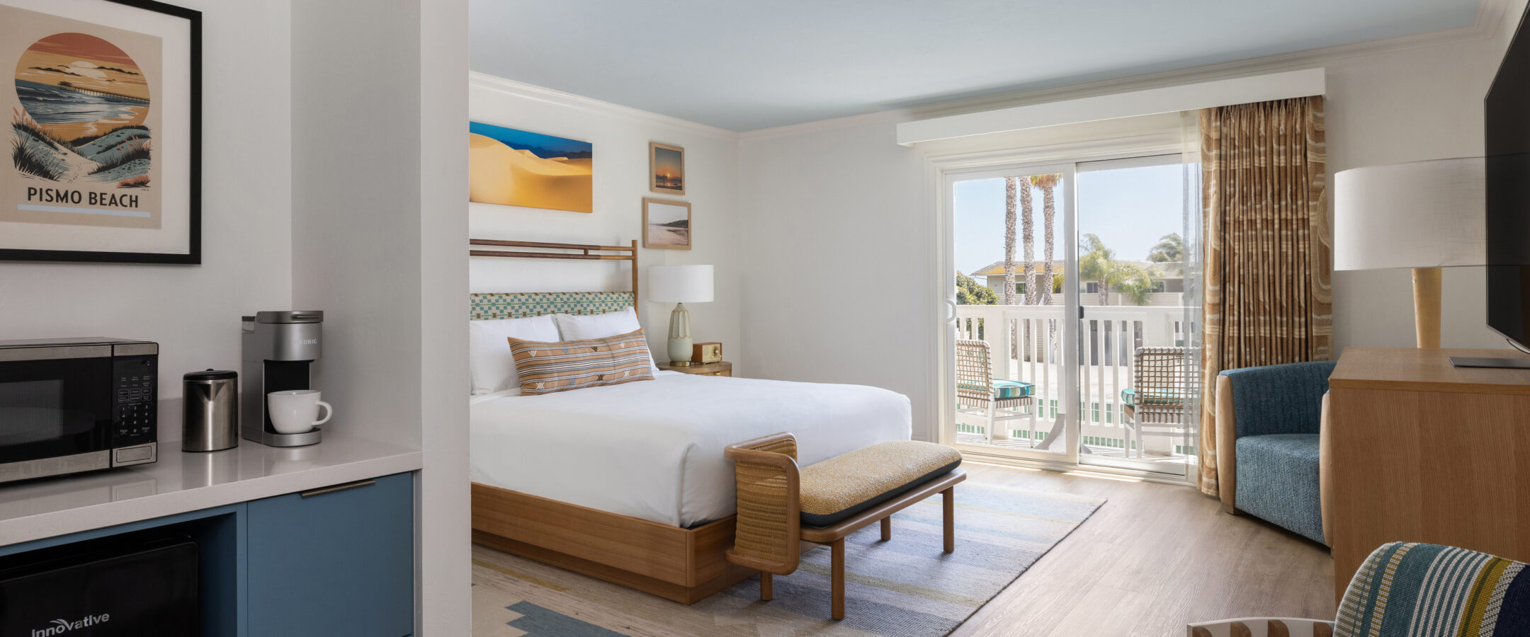 A bright hotel room with a king bed, modern decor, a patterned bench, a seating area, a balcony with outdoor view, mini fridge, microwave, coffee maker, and beach-themed artwork on white walls.