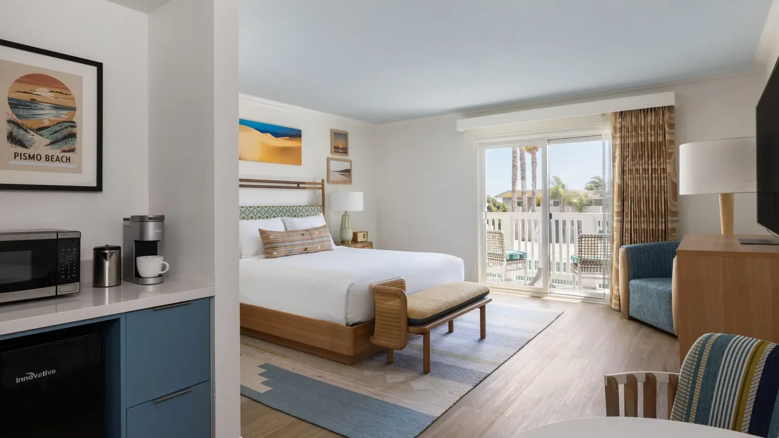A bright hotel room with a king bed, modern decor, a patterned bench, a seating area, a balcony with outdoor view, mini fridge, microwave, coffee maker, and beach-themed artwork on white walls.