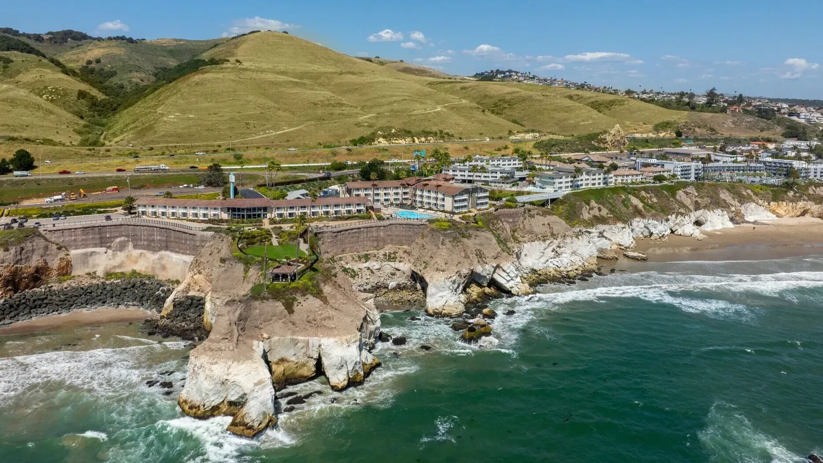 A coastal town with buildings on cliffs overlooking the ocean, green hills in the background, and sandy beaches below invites you to enjoy summer savings as waves crash against the rocky shoreline under a clear blue sky.