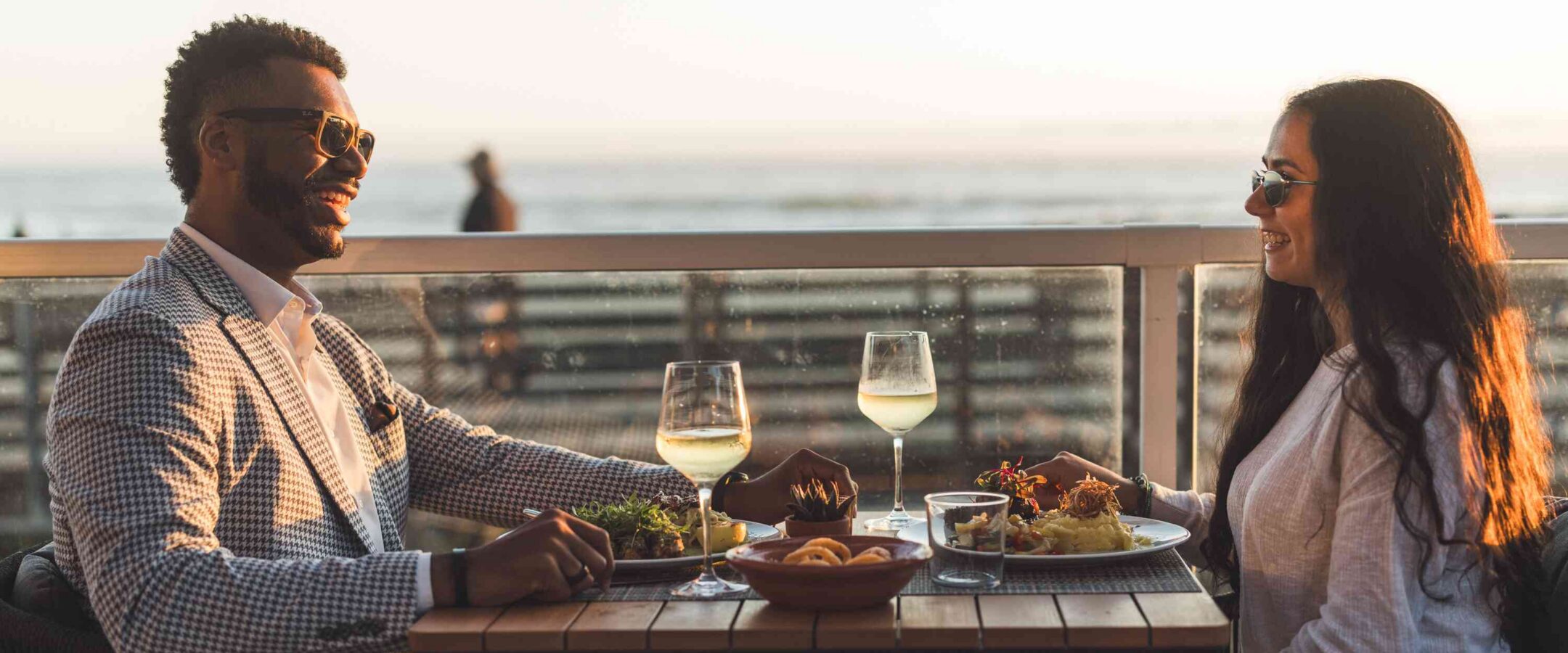 A man and woman wearing sunglasses sit across from each other at an outdoor restaurant table by the ocean, enjoying food and drinks at sunset.