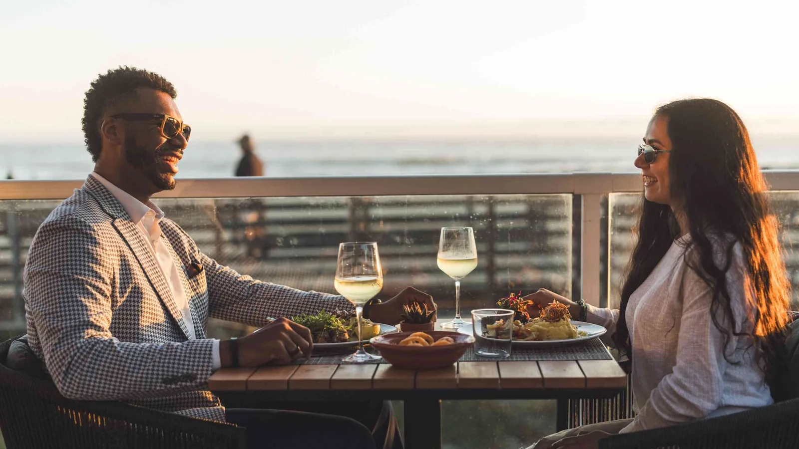 A man and woman wearing sunglasses sit across from each other at an outdoor restaurant table by the ocean, enjoying food and drinks at sunset.