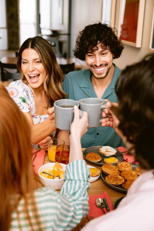 Four friends sit around a breakfast table, smiling and clinking mugs together in a toast. The table is set with pastries, fruit, and drinks, creating a cheerful and lively atmosphere.