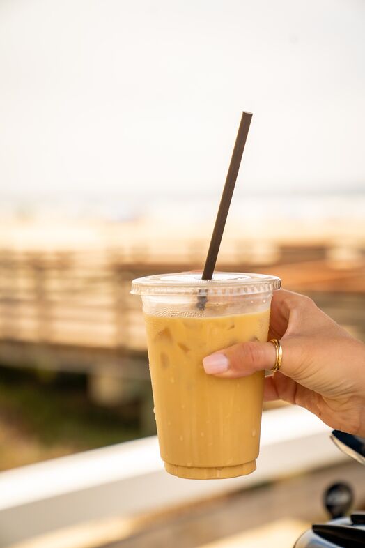 A hand with manicured nails and gold rings holds a plastic cup of iced coffee with a straw, against a blurred outdoor background with warm lighting.