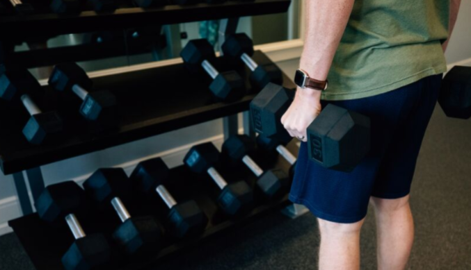 A person in a green shirt and blue shorts holds two dumbbells, standing next to a rack of dumbbells in a gym setting.