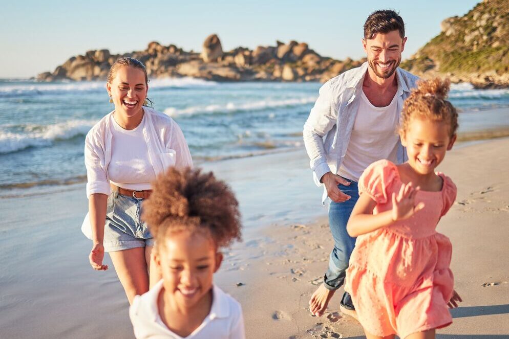 A smiling family of four, two adults and two children, run and laugh together on a sunny beach with waves and rocks in the background.